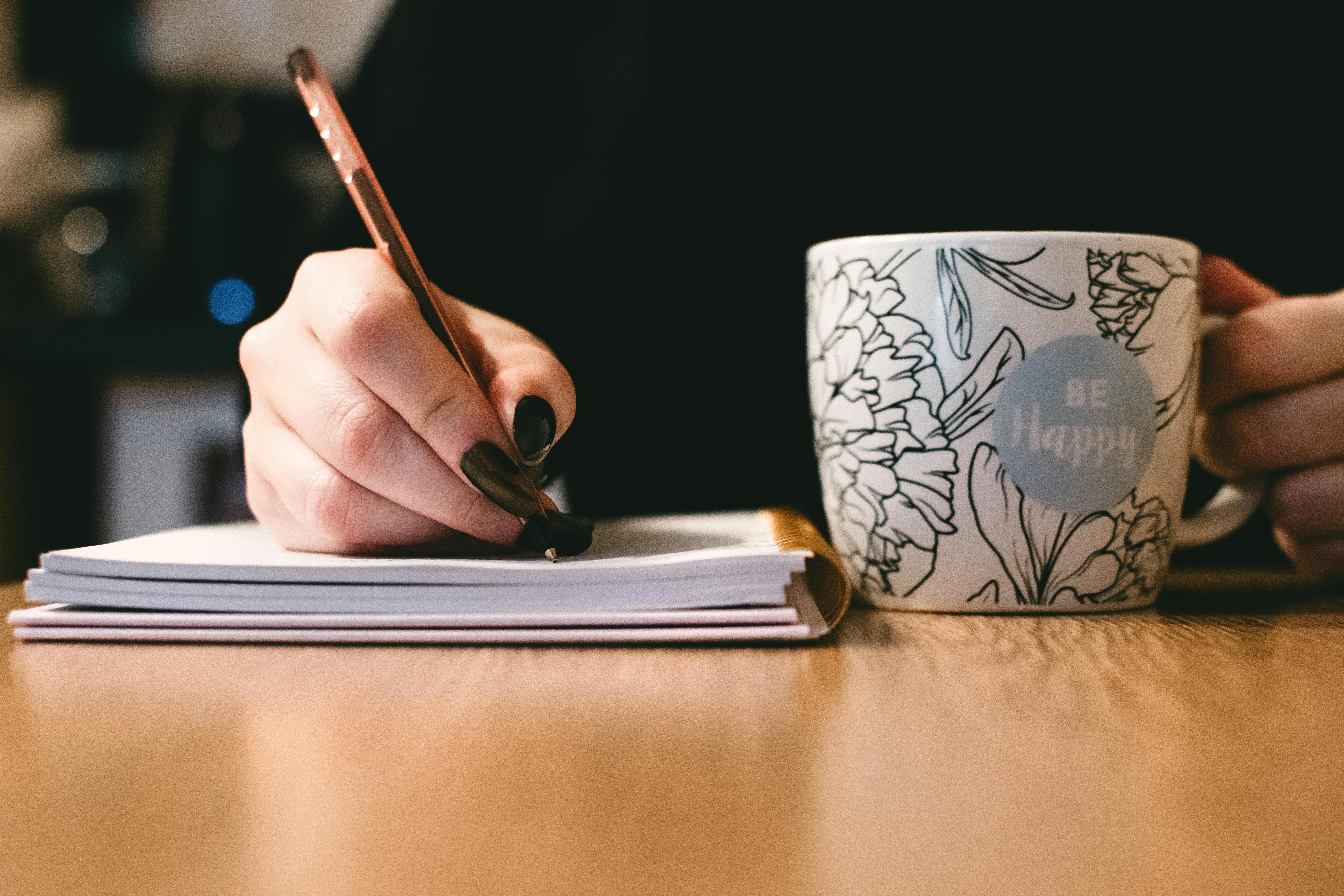women with painted nails writing on notebook and holding a coffee mug